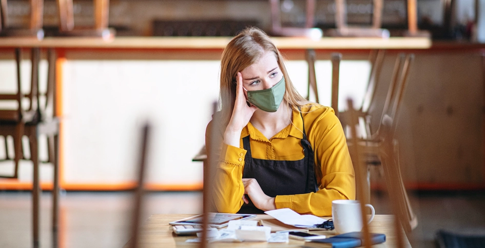 A female business owner with her mask on sitting at a table in an empty restaurant thinking about how covid has impacted her business and will continue to in the future.