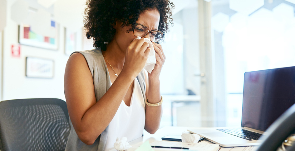 A woman blowing her nose at her desk in the office, building-related illness symptoms can manifest as an illness-related condition but are caused by poor building infrastructure.