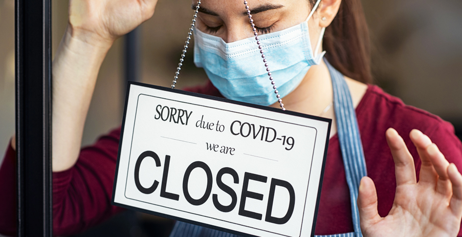 Female business owner leaning against the front door after placing a sign reading, ”Sorry, due to Covid-19 we are closed.” COVID-19 is still somewhat unpredictable and will continue to put pressure on businesses.