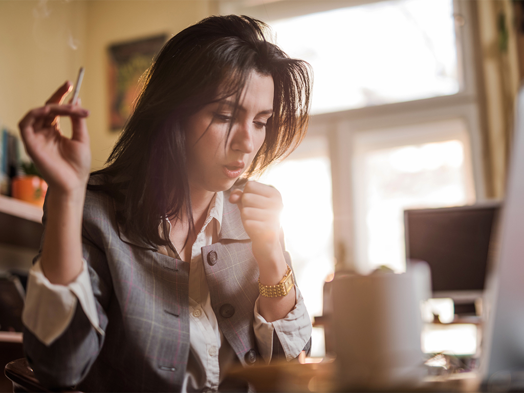 Woman coughing at her desk after smoking. Cigarette smoke contains Benzene which is a volatile organic compound.