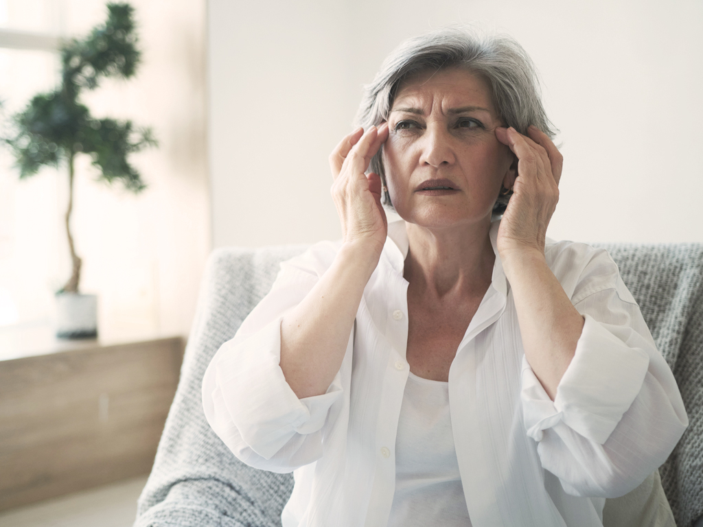 Older woman sitting on a chair rubbing her head due to a migraine.