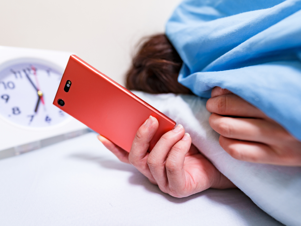 Person hiding under blanket while holding a phone and ignoring wake-up alarm