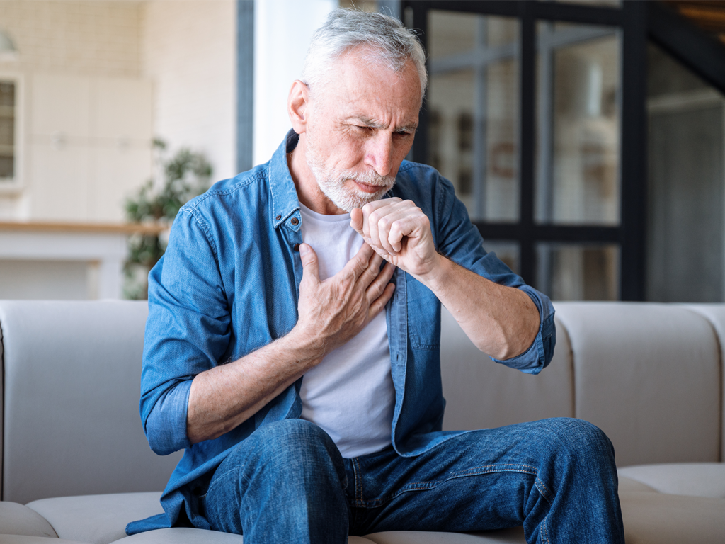 A person sitting on a couch holding his chest, coughing. RSV is an upper respiratory virus that has methods that can help control transmission like ActivePure Technology.