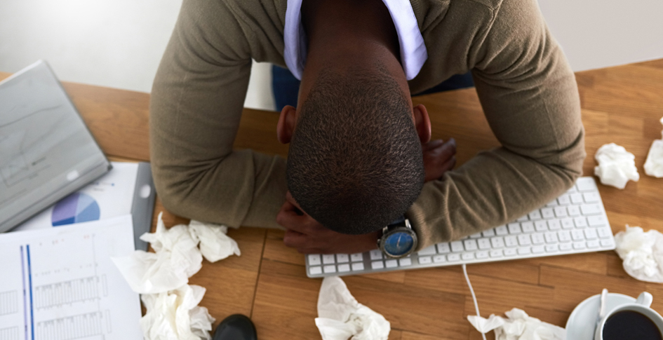 A sick man sitting at his office desk with his head resting on his crossed arms with used tissues surrounding him; sick building syndrome can have the effect of feeling sick by being in a building but cannot be linked to any specific illness.