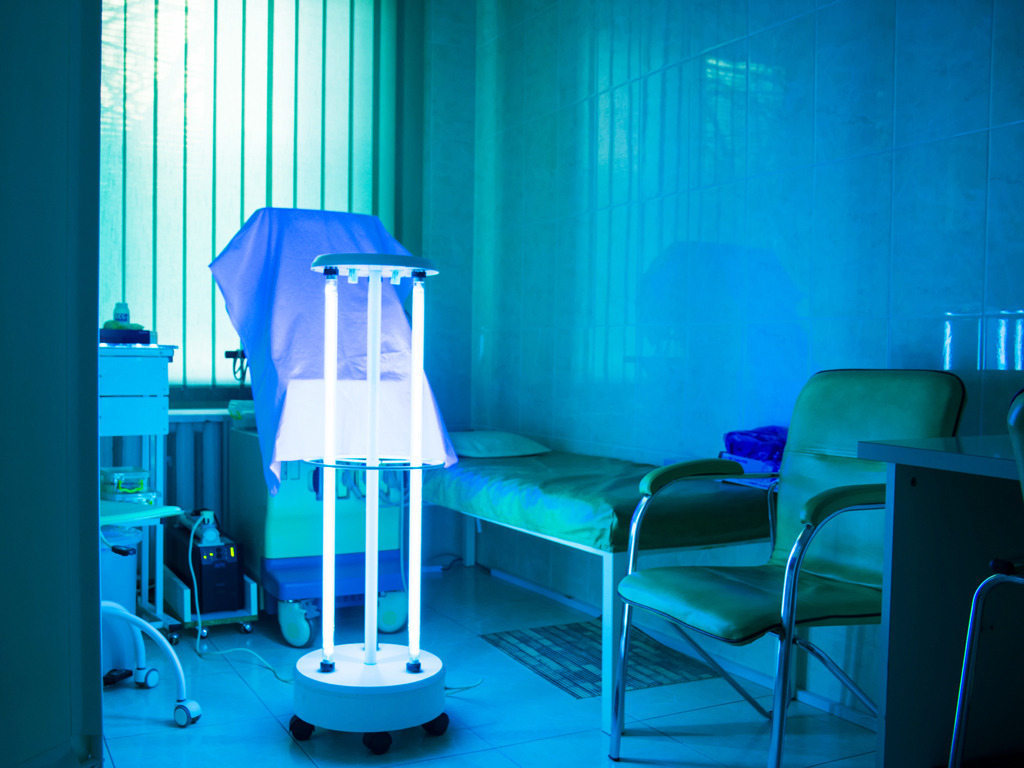 A whole room UV lamp is on in the center of a hospital room. The room must be evacuated while using this unit.