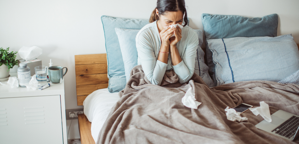 Woman in bed sick with laptop blowing nose with tissues laying in bed during flu season.