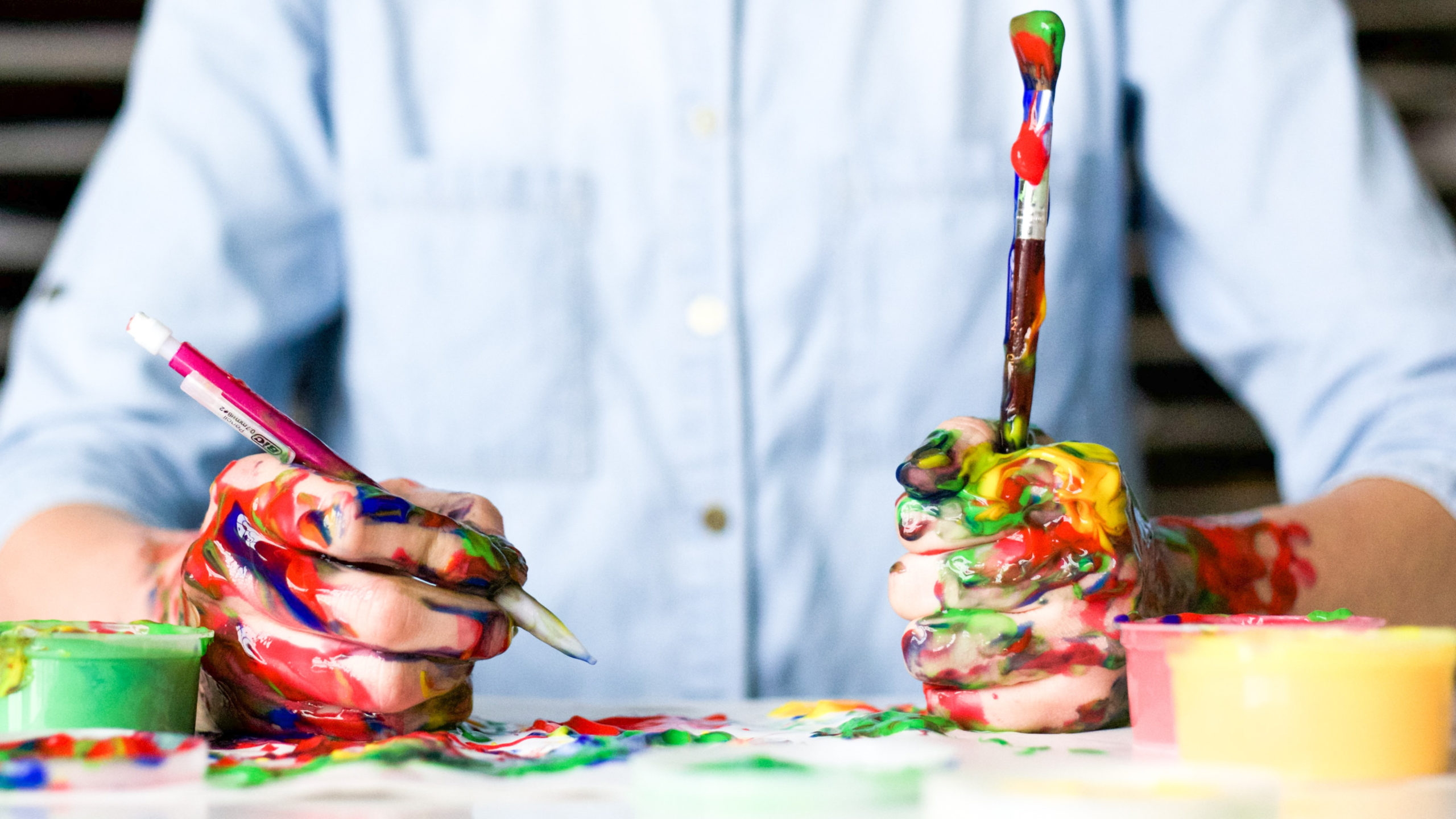 Person with paint covering his hands while holding pencil and paintbrush, paint gives off odors which are volitile organic compounds