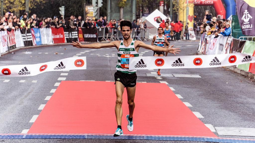 Marathon runner cutting through finish line tape with arms extended, in background are two more runners.